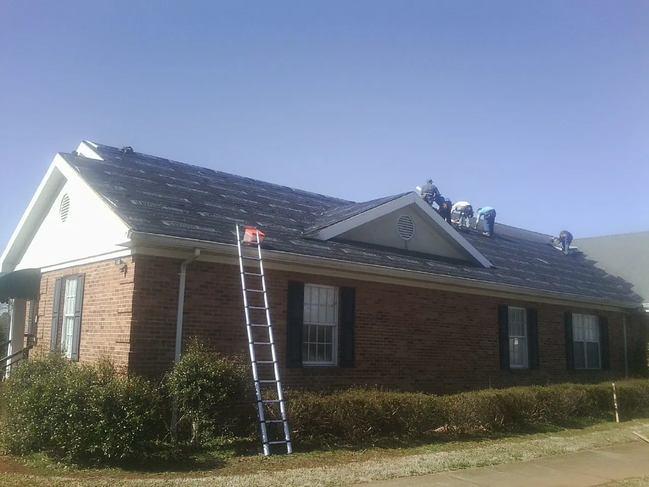 Crew tearing shingles off a brick ranch roof with a ladder set — Metal Roof Repair work by our crew in Sandown