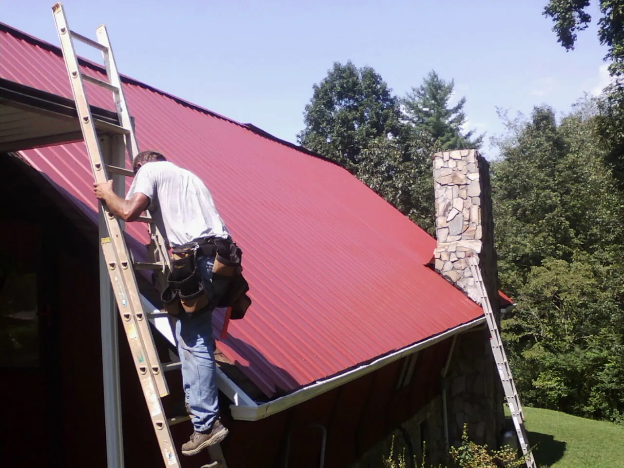 Roofer on a ladder beside a red metal roof and stone chimney — Soffit & Fascia Repair by Roofing Brothers in Sandown